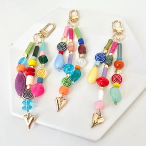 May include: Three colorful beaded keychains with gold-colored hardware and heart charms. Each keychain features a variety of beads in shades of pink, blue, green, yellow, and orange. The keychains are displayed on a white surface.