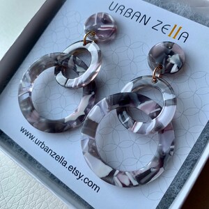 May include: A pair of gray and white patterned resin earrings with gold hoops. The earrings are in the shape of two interlocking circles. The earrings are displayed on a white card with the text "URBAN ZELIA" and "www.urbanzella.etsy.com".