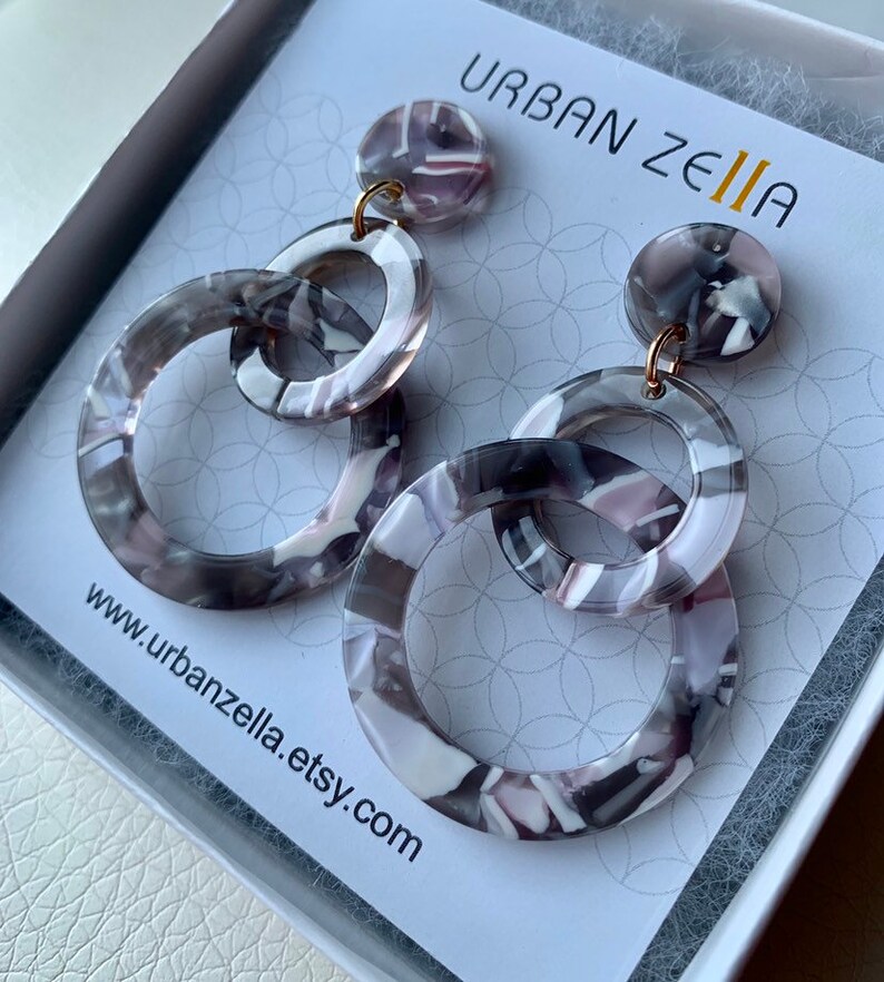 May include: A pair of gray and white patterned resin earrings with gold hoops. The earrings are in the shape of two interlocking circles. The earrings are displayed on a white card with the text "URBAN ZELIA" and "www.urbanzella.etsy.com".