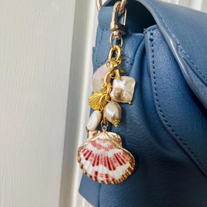 May include: A gold-toned charm with various seashell and pearl accents, attached to a blue handbag. The charm features a red and white seashell, a starfish, and several pearls. The handbag is a solid, medium blue color.