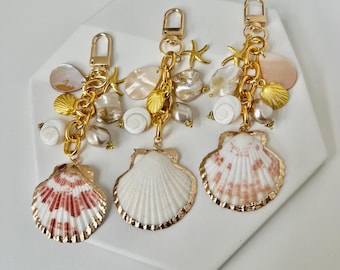 May include: Three seashell keychains with gold-colored hardware. Each keychain features a large shell pendant with gold trim, smaller shells, starfish, and pearl accents. The large shell pendants have white and red patterns.