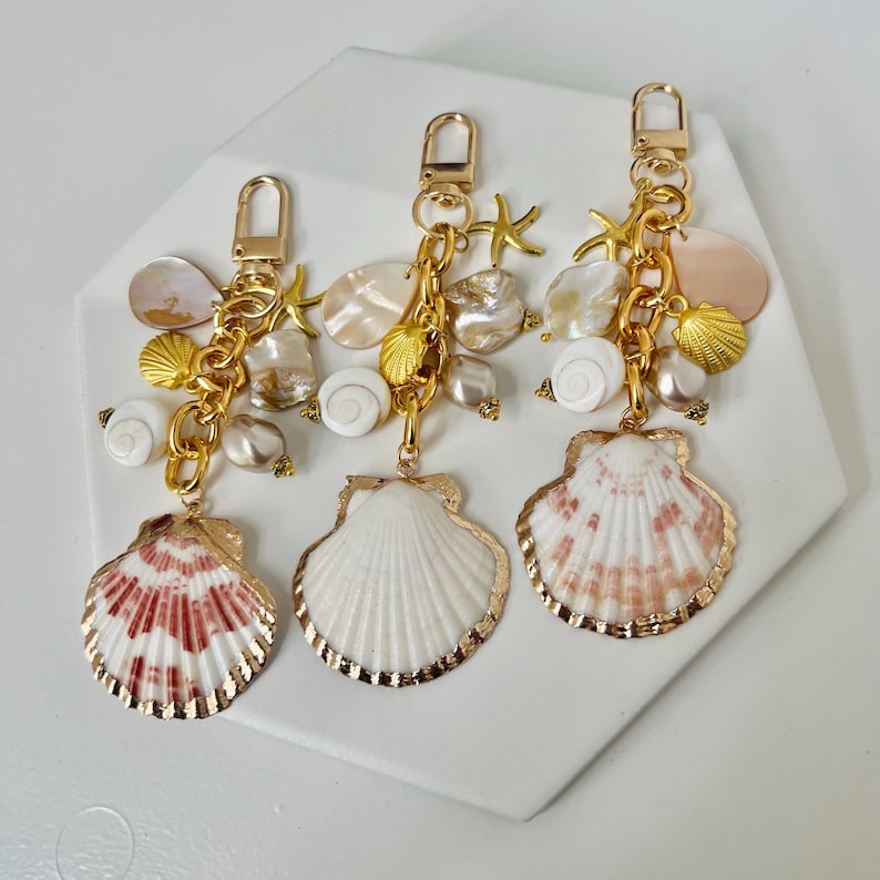 May include: Three seashell keychains with gold-colored hardware. Each keychain features a large shell pendant with gold trim, smaller shells, starfish, and pearl accents. The large shell pendants have white and red patterns.