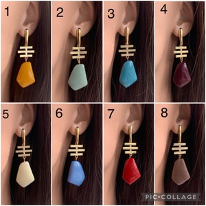 May include: Eight pairs of gold geometric earrings with different colored stone accents. The stones are in shades of yellow, green, blue, red, white, brown, and gray.