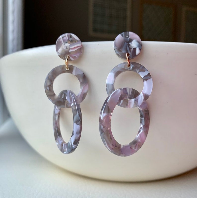 May include: A pair of dangle earrings with a gray and pink marbled pattern. The earrings feature three circles, with the top circle being the smallest and the bottom circle being the largest.
