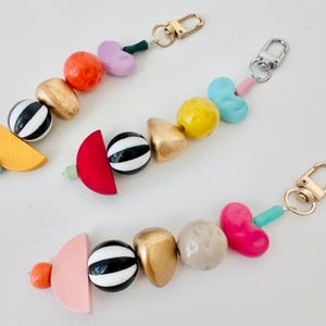 May include: Colorful beaded keychain with gold-tone clasps. The beads are various shapes and colors, including red, pink, gold, black and white striped, and light blue. The keychain is a fun and unique accessory.