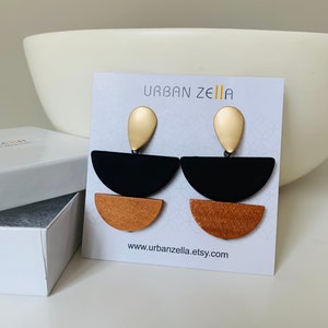 May include: A pair of geometric earrings with black, brown, and gold accents. The earrings feature a teardrop-shaped gold pendant, a black half-circle, and a brown half-circle. The earrings are displayed on a white card with the text "URBAN ZELLIA" and "www.urbanzellia.etsy.com".