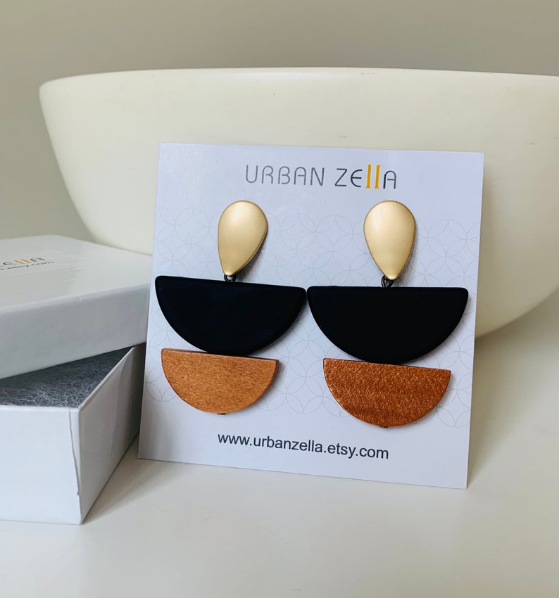 May include: A pair of geometric earrings with black, brown, and gold accents. The earrings feature a teardrop-shaped gold pendant, a black half-circle, and a brown half-circle. The earrings are displayed on a white card with the text "URBAN ZELLIA" and "www.urbanzellia.etsy.com".