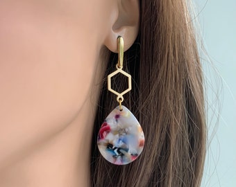 May include: Gold geometric earrings with a teardrop-shaped, multi-colored resin pendant. The pendant features a swirling pattern of white, red, blue, and black.