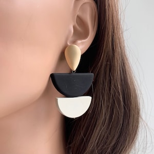 May include: A pair of geometric earrings with black, white, and gold accents. The earrings are made of a lightweight material and feature a teardrop shape at the top, followed by a black half-circle, and a white half-circle.