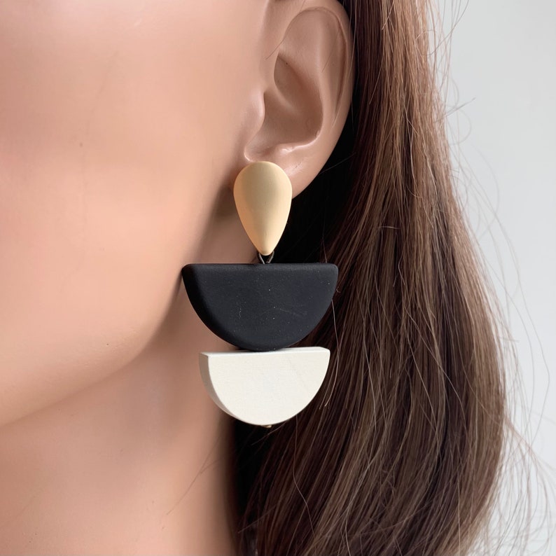 May include: A pair of geometric earrings with black, white, and gold accents. The earrings are made of a lightweight material and feature a teardrop shape at the top, followed by a black half-circle, and a white half-circle.