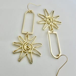 May include: Gold-toned dangle earrings featuring a sun design with a spiral in the center and a rectangular shape. The earrings are made of metal.