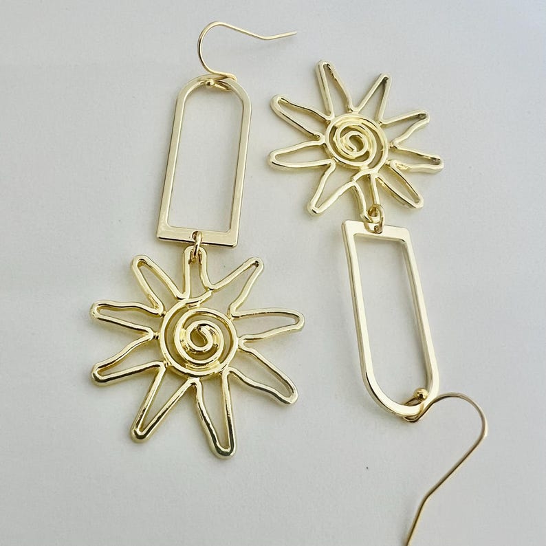 May include: Gold-toned dangle earrings featuring a sun design with a spiral in the center and a rectangular shape. The earrings are made of metal.