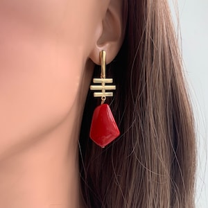 May include: Gold earrings with a red geometric stone pendant. The earrings feature a gold bar design with a geometric shape.