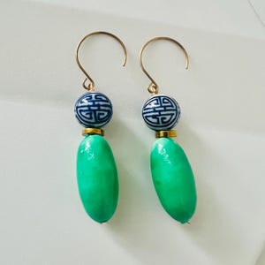 May include: A pair of gold-tone earrings. Each earring features a blue and white patterned bead, a gold-tone accent, and a bright green, oval-shaped bead. The earrings hang from gold-tone ear wires.