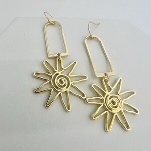 May include: Gold dangle earrings with a geometric rectangle shape and a sun design. The sun has a spiral in the center.