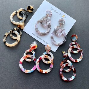 May include: Three pairs of colorful, multi-layered, round, acrylic earrings. The earrings are in various patterns of white, black, pink, blue, and brown. The earrings are displayed on a gray surface with a white card that says "Urban Zella" and a website address.