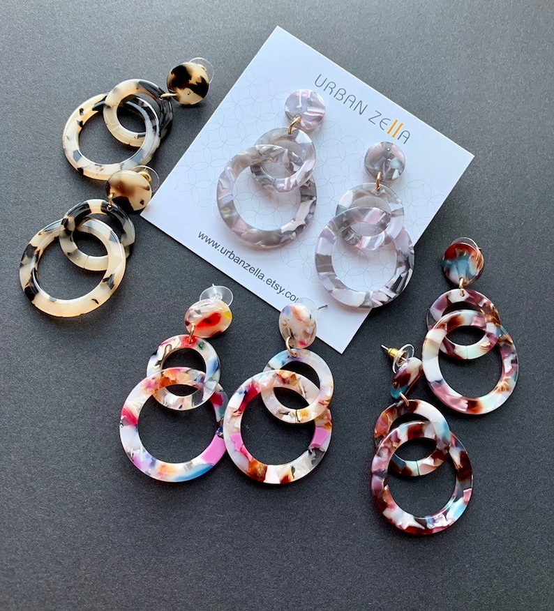 May include: Three pairs of colorful, multi-layered, round, acrylic earrings. The earrings are in various patterns of white, black, pink, blue, and brown. The earrings are displayed on a gray surface with a white card that says "Urban Zella" and a website address.