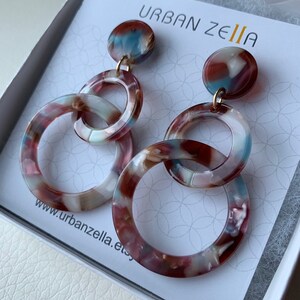 May include: A pair of dangle earrings with three linked circles. The circles are made of a brown, white, and blue marbled plastic. The earrings are hanging from a gold hook.