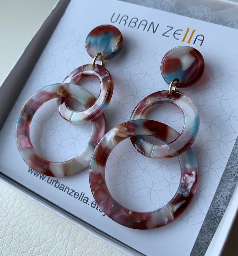 May include: A pair of dangle earrings with three linked circles. The circles are made of a brown, white, and blue marbled plastic. The earrings are hanging from a gold hook.