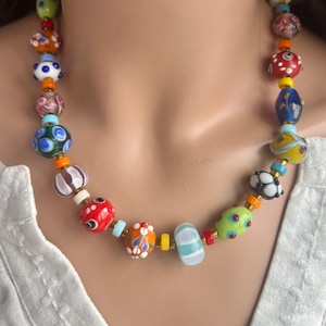 Könnte beinhalten: Eine farbenfrohe Perlenkette mit einer Vielzahl von Glasperlen in Rot, Blau, Grün, Orange und Weiß. Die Perlen sind in verschiedenen Formen und Größen gehalten und mit goldfarbenen Metallakzenten aufgereiht. Ein lebendiges Accessoire.