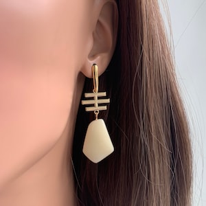 May include: Gold earrings with a white geometric pendant. The pendant is shaped like a teardrop and has a gold bar design above it.