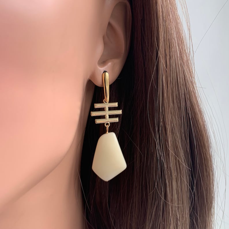 May include: Gold earrings with a white geometric pendant. The pendant is shaped like a teardrop and has a gold bar design above it.