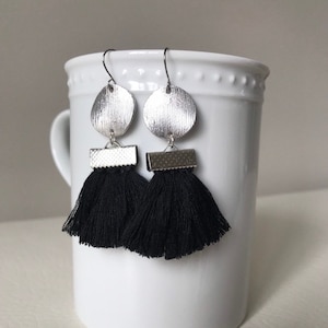 May include: Silver earrings with black tassel fringe. The earrings feature a round, textured silver disc and a silver bar.