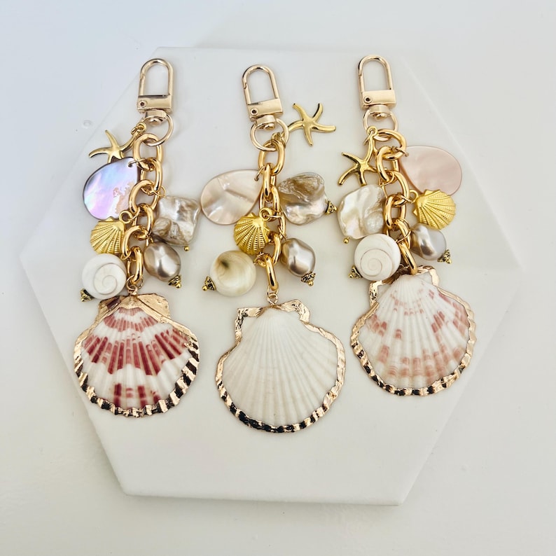May include: Three seashell keychains with gold-colored hardware. Each keychain features a large seashell pendant with a gold trim, along with smaller shells, pearls, and starfish charms. The seashells have white and pink hues.