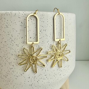 May include: Gold dangle earrings with a rectangular frame and a sun-shaped charm.