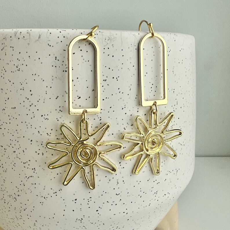 May include: Gold dangle earrings with a rectangular frame and a sun-shaped charm.