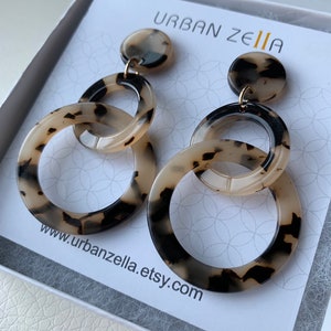 May include: A pair of tortoise shell patterned earrings with three interlocking circles. The earrings are hanging from a small round disc. The earrings are in a white box with the text "URBAN ZELIA" and "www.urbanzella.etsy.com".