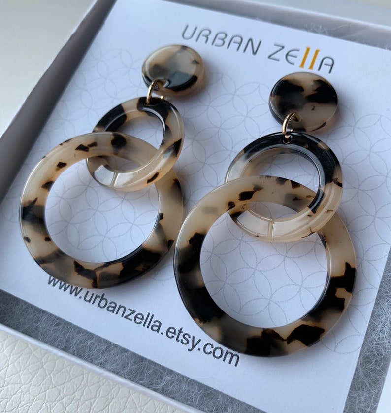 May include: A pair of tortoise shell patterned earrings with three interlocking circles. The earrings are hanging from a small round disc. The earrings are in a white box with the text "URBAN ZELIA" and "www.urbanzella.etsy.com".