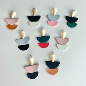 May include: Set of 10 pairs of geometric earrings. Each earring features three stacked half-circles in different colors, including pink, red, green, black, white, and brown. The earrings have a gold-colored teardrop-shaped top.