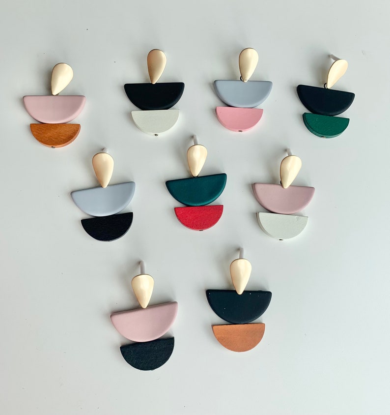 May include: Set of 10 pairs of geometric earrings. Each earring features three stacked half-circles in different colors, including pink, red, green, black, white, and brown. The earrings have a gold-colored teardrop-shaped top.