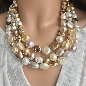 May include: A multi-strand necklace featuring irregularly shaped pearls in shades of white, gold, and blush. The necklace has gold-toned accents and is worn against a white blouse.