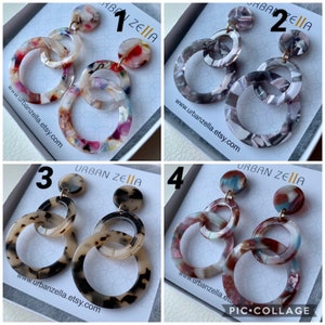 May include: Four pairs of multi-colored, layered, round, acrylic hoop earrings. Each pair is unique with different color combinations and patterns. The earrings are displayed on a white background with the text "URBAN ZELIA" and a website address.