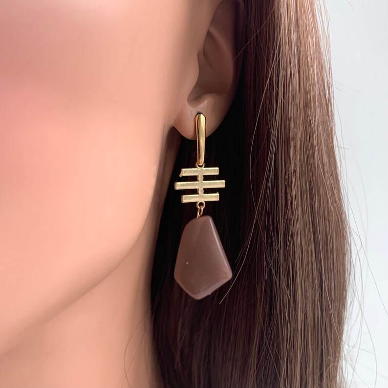 May include: Gold geometric earrings with a brown stone pendant. The earrings have a simple, modern design.