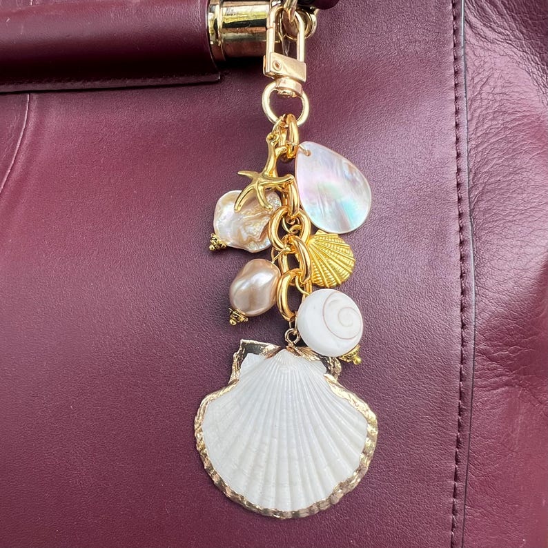 May include: A gold-toned seashell charm with a white shell and gold trim, hanging from a gold chain. The charm features various seashell and pearl accents, including a starfish and iridescent teardrop-shaped charm. The background is a burgundy leather surface.