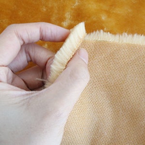 May include: A close-up of a light brown faux fur fabric with a soft, fuzzy texture. The fabric is being held in a hand, revealing a woven backing.