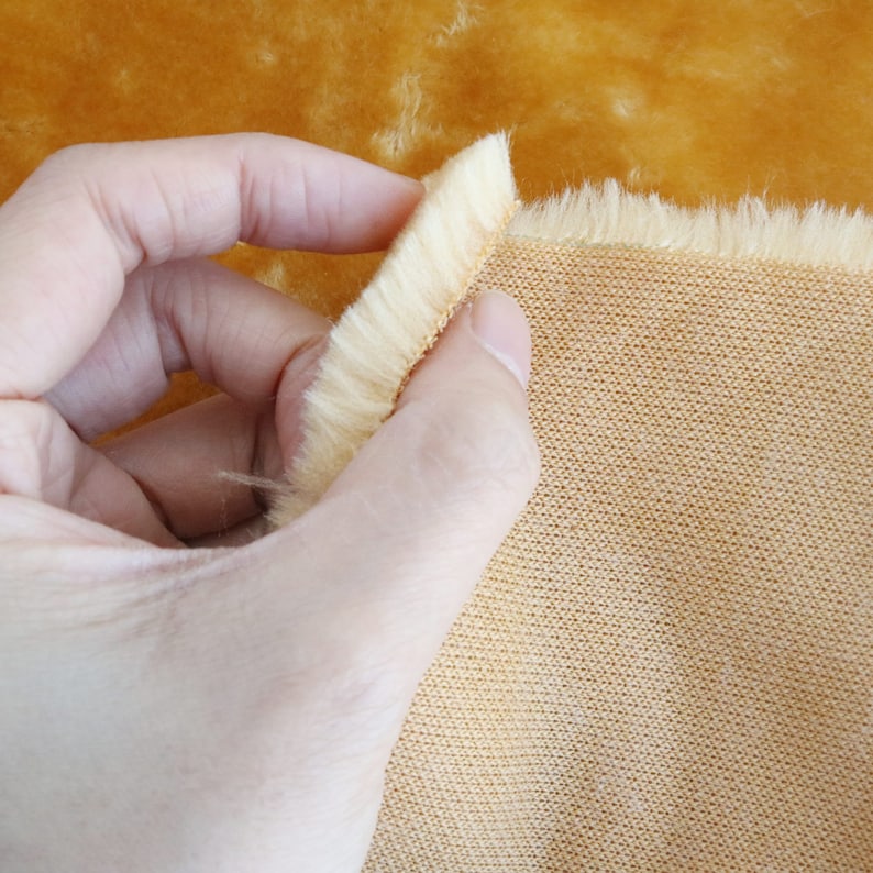 May include: A close-up of a light brown faux fur fabric with a soft, fuzzy texture. The fabric is being held in a hand, revealing a woven backing.