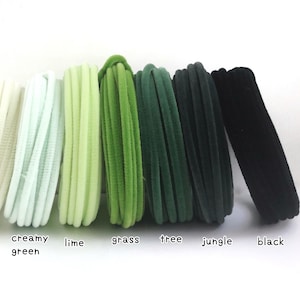 May include: A collection of six elastic hair ties arranged in a row, featuring a gradient of green shades labeled creamy green, lime, grass, tree, jungle, and black. Each hair tie has a soft, velvet-like texture and a circular, looped design.