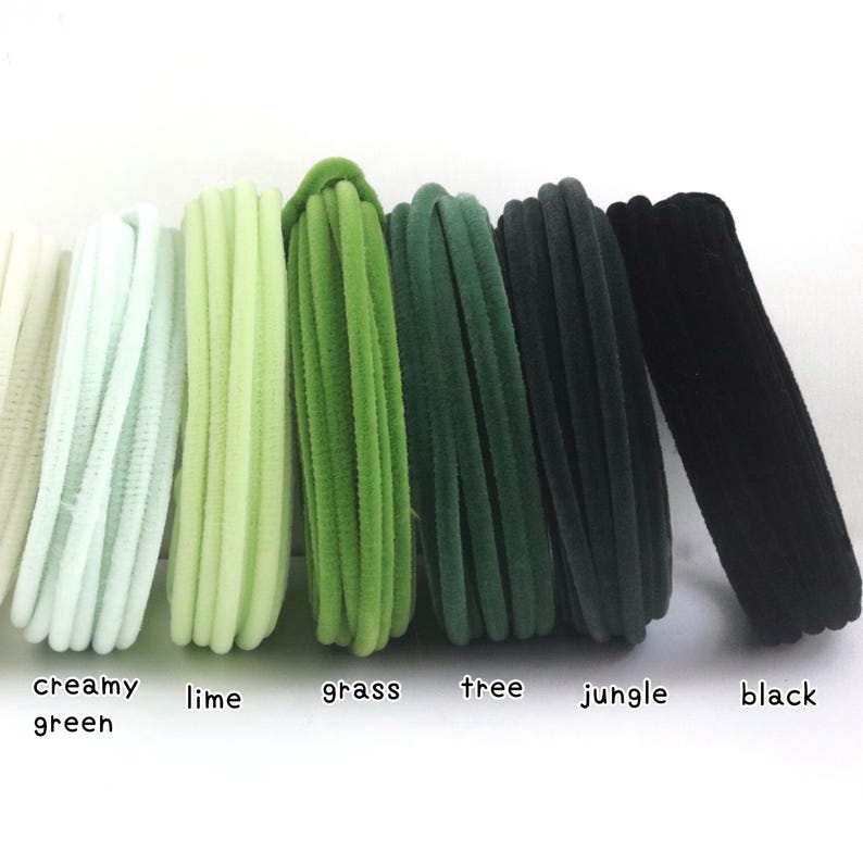 May include: A collection of six elastic hair ties arranged in a row, featuring a gradient of green shades labeled creamy green, lime, grass, tree, jungle, and black. Each hair tie has a soft, velvet-like texture and a circular, looped design.