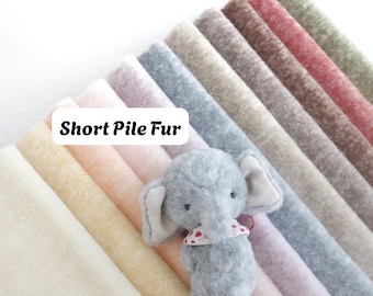 Puede incluir: Una colección de muestras de tela de piel de pelo corto en varios colores, incluyendo crema, beige, rosa, gris, marrón, rojo y verde, dispuestas en abanico con un pequeño elefante de peluche gris que lleva un pañuelo blanco con corazones.