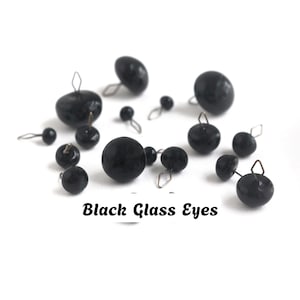 Könnte beinhalten: Eine Sammlung verschieden großer schwarzer Glasaugen mit Drahtschlaufen zur Befestigung. Jedes Stück ist glatt, rund und glänzend. Der Text Black Glass Eyes ist unten im Bild in Schwarz aufgedruckt und identifiziert diese Bastelkomponenten.