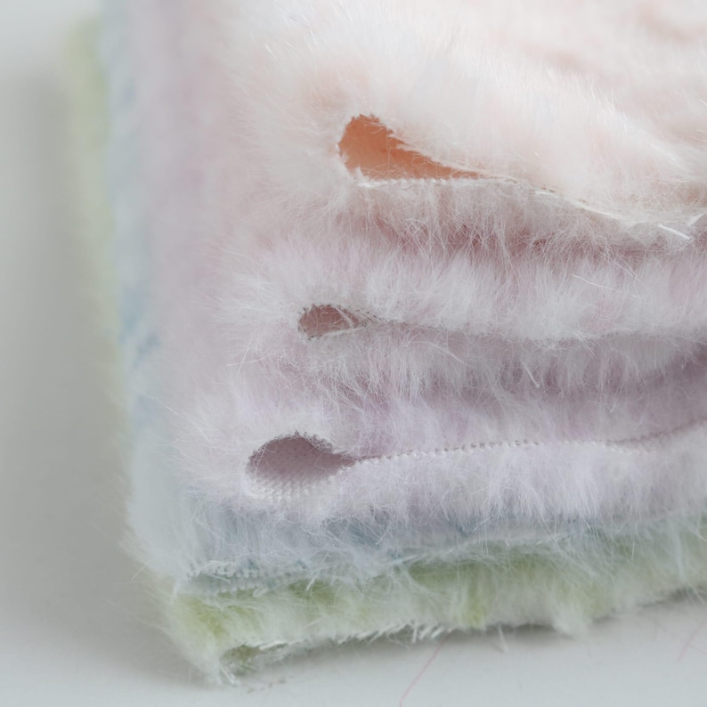 May include: Close-up of a stack of fluffy, pastel-colored fabric. The fabric has a soft, fuzzy texture and is layered in shades of pink, blue, and green. There are several oval-shaped cutouts visible in the fabric.