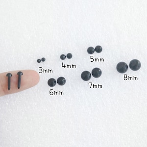 Peut inclure: Assortiment d'yeux de s&eacute;curit&eacute; en plastique noir pour l'artisanat, allant de 3 mm &agrave; 8 mm. Les yeux sont pr&eacute;sent&eacute;s sur une surface blanche avec un doigt tenant deux yeux plus petits.