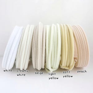 May include: A set of six circular hair ties arranged in a row, featuring a ribbed texture. The colors from left to right are white, creamy white, beige, light yellow, soft yellow, and peach. Each hair tie is labeled with its respective color name.