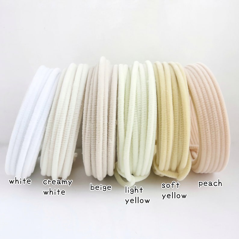 May include: A set of six circular hair ties arranged in a row, featuring a ribbed texture. The colors from left to right are white, creamy white, beige, light yellow, soft yellow, and peach. Each hair tie is labeled with its respective color name.