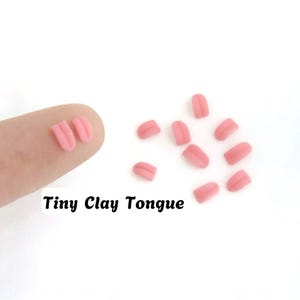 Puede incluir: Una colección de formas de lengua de arcilla rosa en miniatura, cada una con una hendidura vertical central. Las piezas pequeñas, lisas y con forma de lengua están dispersas por el encuadre, con dos piezas adicionales sobre una yema de dedo. El texto Tiny Clay Tongue es visible en fuente negra.