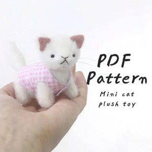 Op de afbeelding: Een handgemaakt wit pluche kattenknuffel met een roze ruitjeshemd en roze oorpunten. De kat heeft een gestikt gezicht en snorharen. De tekst op de afbeelding luidt "PDF Pattern Mini cat plush toy."
